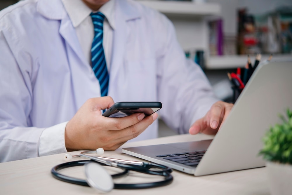A doctor in a white coat using a smartphone and laptop at a desk, with a stethoscope nearby.